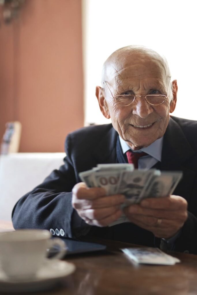 Confident senior businessman holding money in hands while sitting at table near laptop